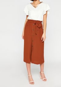 Top bianco con maniche a volant e scollo a V con accenti dorati, abbinato a una gonna arancione al ginocchio con bottoni e cintura annodata.