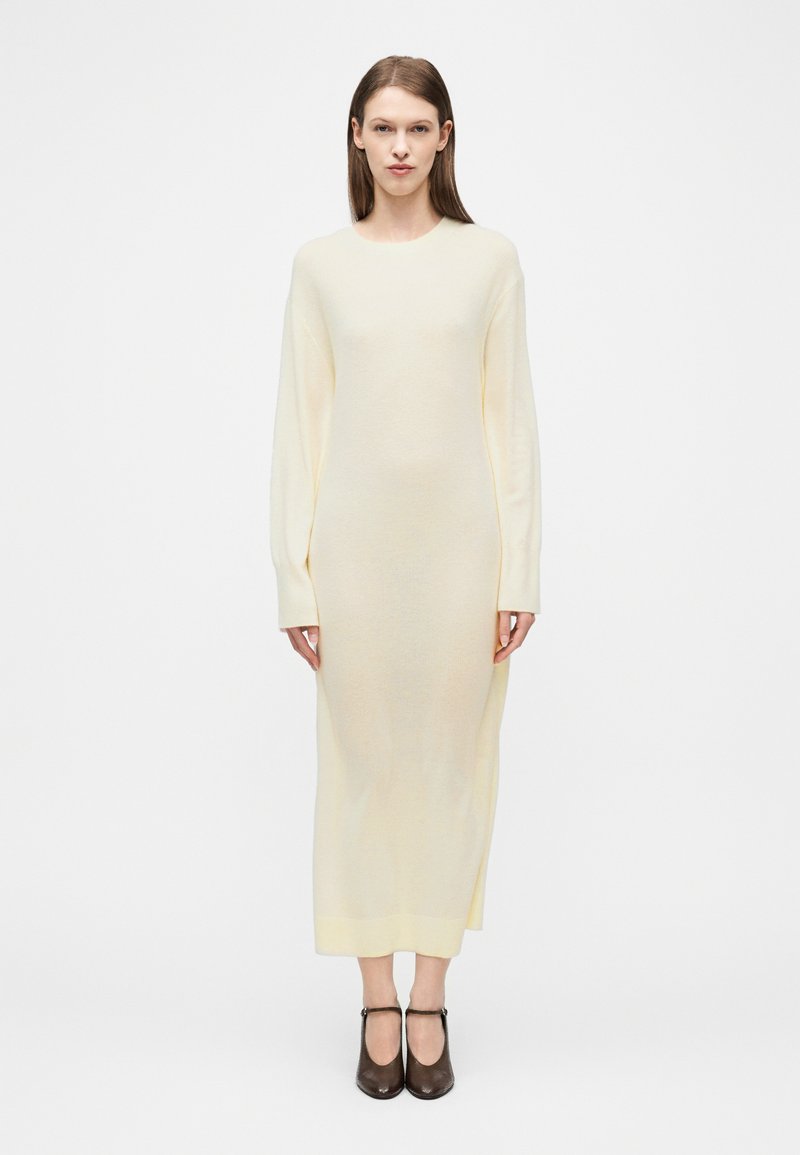 Γυναίκα στέκεται μπροστά σε μονόχρωμο φόντο, φορώντας ένα πλεκτό φόρεμα εκρού με μακριά μανίκια μέχρι τον αστράγαλο και σκούρα καφέ παπούτσια Mary Jane.