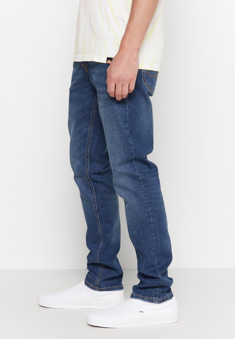 Jeans de mezclilla azul oscuro con corte slim, que cuentan con un diseño clásico de cinco bolsillos, costuras visibles y un efecto desgastado cerca de las rodillas.