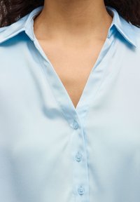 Ανοιχτό μπλε μπλούζα με κουμπιά εμπρός, V-λαιμόκοψη και γιακά. Λείο ύφασμα με απαλή γυαλάδα, έξι στρογγυλά κουμπιά ορατά.