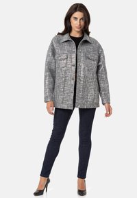 Grau strukturierte Jacke mit zwei Brusttaschen, Knopfverschluss und weitem Kragen, kombiniert mit dunklen Skinny-Jeans und schwarzen High Heels.