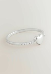 Srebrny pierścionek z sercem jako centralnym elementem i małymi, przejrzystymi kamieniami na cienkiej opasce na jednolitym jasnym tle.