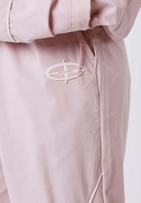Chaqueta de color rosa claro con un bolsillo lateral, presenta un logo ovalado PXP en blanco y rosa y un cordón ajustable cerca de la apertura del bolsillo.