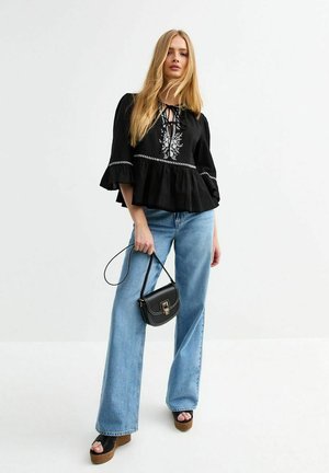 Blouse noire brodée avec des manches larges et évasées, ceinturée à la taille, associée à un jean large bleu clair et un petit sac à main noir.