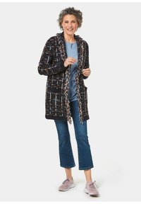 Schwarzer und mehrfarbiger langer Cardigan mit Fransenbesatz, Taschen; getragen über einem blauen Shirt und kombiniert mit cropped Jeans und hellen Schuhen.