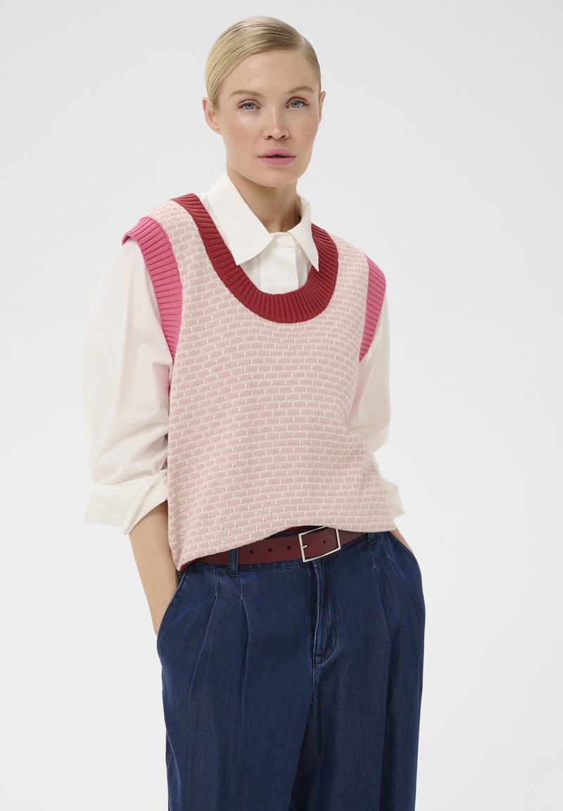 Femme aux cheveux blonds portant un gilet tricoté sans manches rose sur une chemise blanche à col et un pantalon bleu taille haute, les mains dans les poches.