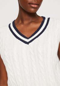 Bílý pletený bezrukávový svetr s výstřihem do V, s námořnickými a bílými pruhovanými ribovými pruhy. Textura vykazuje vzory v kabelovém úpletu.