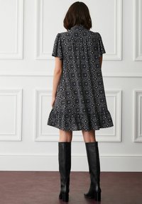 Kvinde iført en mørk mønstret kortærmet kjole med en flæsekant, parret med sorte knæhøje hæle støvler, stående mod hvide panelvægge.