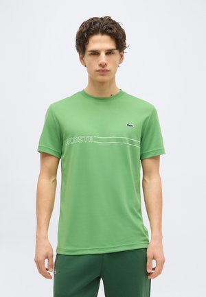 Νεαρός άνδρας στέκεται, φορώντας πράσινο t-shirt Lacoste με λογότυπο και κείμενο, συνδυασμένο με σκούρο πράσινο παντελόνι, απλό φωτεινό φόντο.