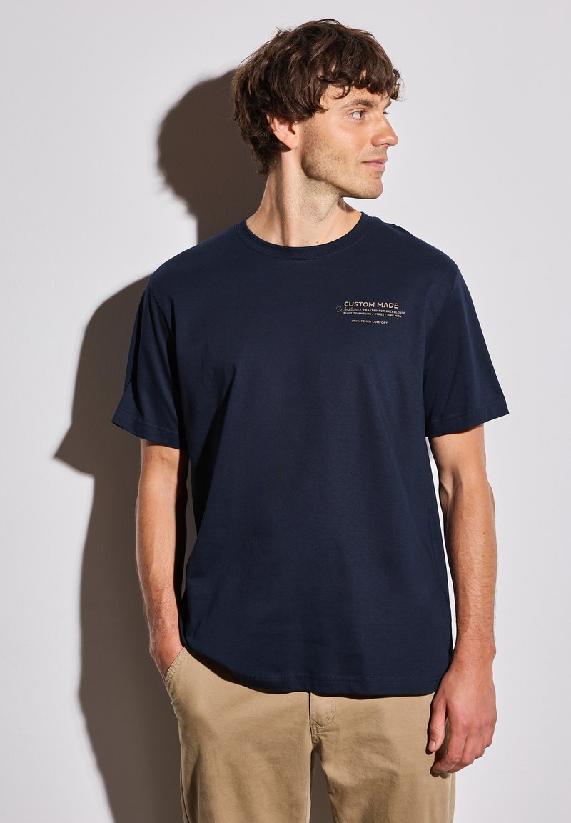 Mörkblå bomullströja med kort ärm, avslappnad passform, har vit text "CUSTOM MADE" och detaljtryck på bröstet.