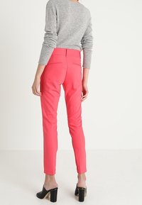 Kirkkaita vaaleanpunaisia, istuvia housuja, jotka on valmistettu joustavasta materiaalista. Niissä on takataskut ja sileä pinta, yhdistettynä mustiin korkokenkiin.