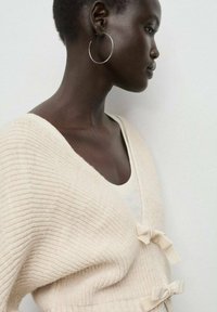Cardigan color crema, a coste, con scollatura a V profonda, dotato di due piccole decorazioni a fiocco nella parte anteriore e maniche lunghe.