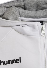 Haut à capuche blanc avec un intérieur en gris, doté d'une fermeture éclair à l'avant et d'un texte noir en logo. Le tissu semble lisse avec une texture douce.