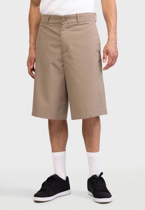 Άνδρας που φοράει μπεζ σορτς μέχρι το γόνατο, λευκές κάλτσες έως τον αστράγαλο και μαύρα αθλητικά παπούτσια, στέκεται μπροστά σε λευκό απλό φόντο.