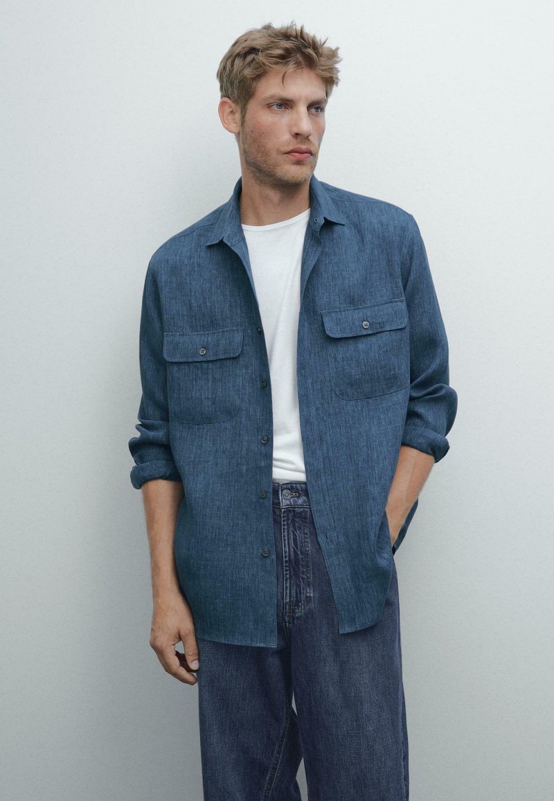 Giovane uomo che indossa una camicia azzurra con bottoni sopra una maglietta bianca e dei pantaloni jeans scuri larghi, in piedi con una mano in tasca su uno sfondo neutro.