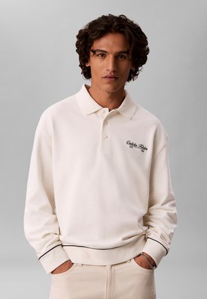 Giovane uomo con capelli ricci che indossa una polo bianca a maniche lunghe e pantaloni chiari, mani nelle tasche, sfondo grigio neutro.