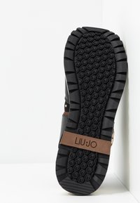 Scarpa nera con suola in gomma, caratterizzata da una texture sul battistrada, un accento in pelle marrone con logo e una combinazione di materiali lisci e scamosciati.