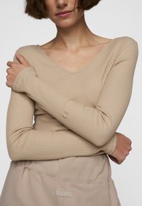 Beige geripptes Langarmoberteil mit V-Ausschnitt und enganliegenden Ärmeln, kombiniert mit einer dazu passenden Unterbekleidung mit einem Logo-Tag.