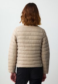 Giubbotto imbottito di colore beige chiaro, caratterizzato da un design a righe orizzontali e un collo rotondo. Texture morbida con hardware minimo.