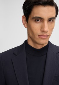 Blazer blu navy con sottili righe verticali, abbinato a un dolcevita scuro aderente. Texture morbida, design su misura e linee pulite.