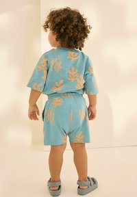 Türkisfarbenes zweiteiliges Outfit mit kurzen Ärmeln und Shorts, verziert mit orangen Blattmustern. Der Stoff wirkt weich und ist auf Komfort ausgelegt.