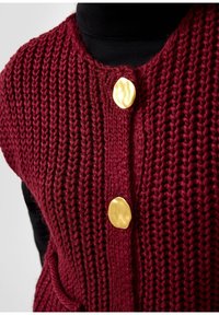 Bordo adīts bezmugura žaketes ar teksturētu apdari, kas aprīkota ar trim lielām zelta apaļām pogām un divām priekšējām kabatām. Melna golfa kreklā zem tā.