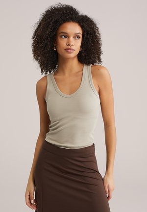 Lokkis juustega naine, kes kannab beežikat varrukateta topi ja kõrge vöökohaga pruuni seelikut, seisab ühevärvilise heleda tausta ees.