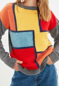 Bunter Strickpullover mit geometrischen Mustern in Rot, Gelb, Blau und Orange. Graue Ärmel und gerippte Bündchen verleihen Textur.