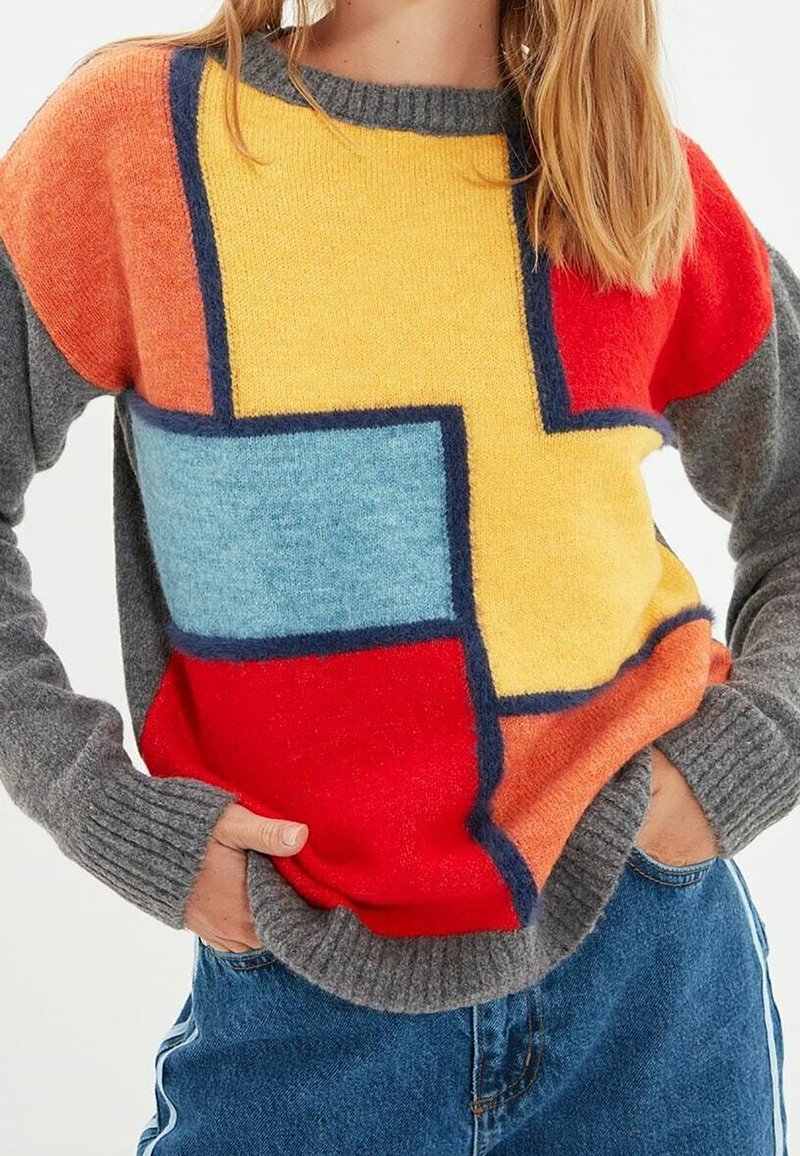 Bunter Strickpullover mit geometrischen Mustern in Rot, Gelb, Blau und Orange. Graue Ärmel und gerippte Bündchen verleihen Textur.