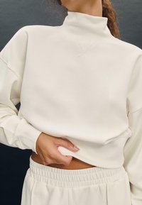 Felpa girocollo di colore crema con colletto alto, vestibilità morbida, texture soffice e polsini a coste, abbinata a pantaloni con vita elastica dello stesso colore.