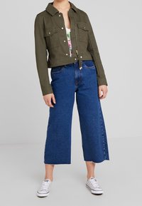 Oliwkowa krótka kurtka z przednimi kieszeniami i złotym zamkiem, zestawiona z niebieskimi dżinsami o szerokich nogawkach i białymi trampkami.