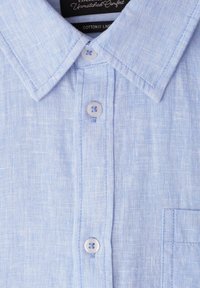 Chemise bleu clair en mélange de coton et lin avec col boutonné, trois boutons blancs et coutures visibles sur la poche avant.