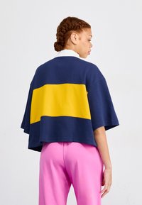 Navy and yellow color-blocked oversized top with short sleeves, featuring a white collar. Paired with pink pants, smooth fabric texture.