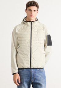 Light beige padded jacket with a hood, featuring black zippers, ribbed cuffs, and a zippered pocket on the left sleeve.