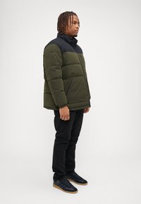 Veste en duvet verte et noire, en matériau épais avec un motif matelassé, poches zippées à l'avant, portée sur un pantalon noir et des baskets noires.