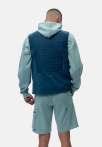 Tamsiai mėlynas siuvinėtas liemenė su minkštu žaliu džemperiu po juo, turi užtrauktuką kišenėje ant šortų ir reibotus rankogalius patogumui.