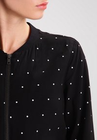 Giacca bomber nera con chiusura a zip, collo rotondo e pois bianchi. Realizzata in materiale leggero con una texture liscia.