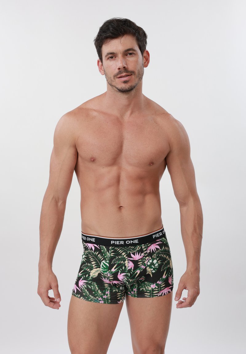 Skjorteløs mand stående foran en hvid baggrund iført sorte boxershorts med grønne og pink tropiske bladmønstre.