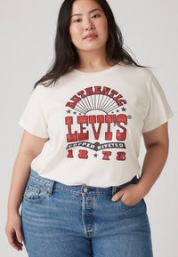 Kremo spalvos trumpų rankovių marškinėliai su ryškiu raudonu ir juodu retro logotipu, papuoštu žvaigždėmis ir saulės spinduliais. Pora su aukštu liemeniu mėlynomis džinsomis.