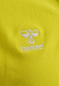 Weißes, besticktes Hummel-Bienenlogo und Text auf leuchtend gelbem, strukturiertem Stoffhintergrund.