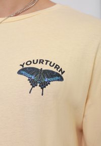 Geltona medvilninė marškinė su juodąją drugelio grafika, turinčia iriškančių mėlynų akcentų, ir žodžiu "YOURTURN" virš piešinio.