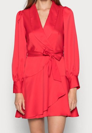 Frau trägt ein rotes, langärmliges Wickelkleid mit einer gebundenen Taillenschleife und einem gerafften Saum vor weißem Hintergrund.