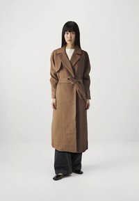 Sieviete stāv, tērpusies garā brūnā jostotā mētelī, uz melnām platām biksēm un melniem norādītajiem apaviem uz vienkārša baltā fona.