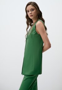 Gilet verde senza maniche con taglio diritto, caratterizzato da una texture liscia e un design minimalista, abbinato a pantaloni a sigaretta coordinati.