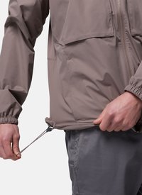 Personne ajustant le cordon de serrage à l'ourlet d'une veste mauve zippée avec des poignets élastiques, portant un pantalon gris.