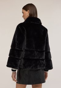 Kobieta z prostymi, brązowymi włosami nosi czarną kurtkę z sztucznego futra i czarną skórzaną spódnicę, pokazana od tyłu na jednolitym tle.