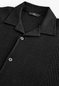 Schwarzes Poloshirt mit strukturiertem Design, flachem Kragen und Knopfschluss mit grauen Knöpfen. Hergestellt aus einem gemusterten Stoff.