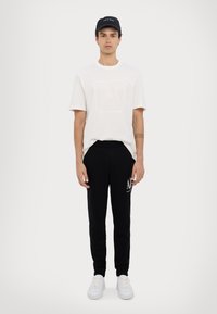 Weißes Baumwoll-T-Shirt mit Logo, schwarze Jogginghose mit weißem Text und weiße Sneaker. Schlichte, lockere Passform, stehend vor einem schlichten Hintergrund.