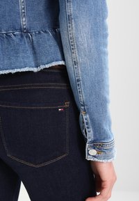 Jeansjacka med fransad nederkant, ljusblå färg och knäppt manschett, i kombination med mörkblå jeans som har en röd och vit detalj på fickan.
