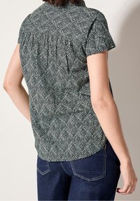 Korte mouwen blouse in zwart en crème met bladpatroon, geplooide achterkant en afgeronde zoom. Gecombineerd met donkere denim jeans.